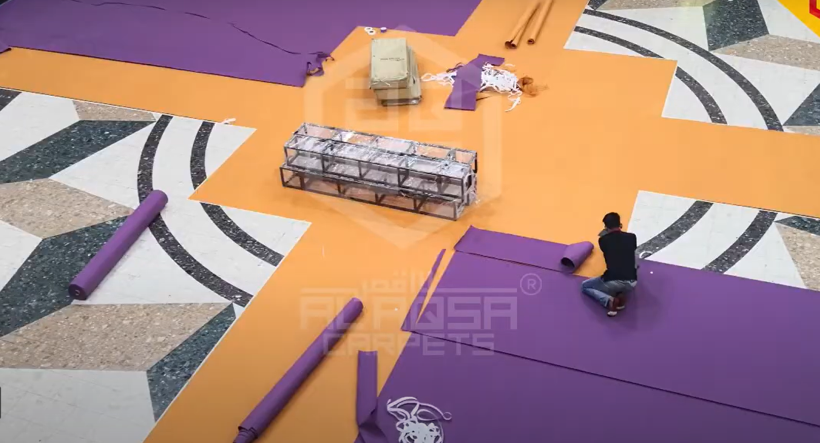 Worker installing large purple and orange exhibition event carpet sections over a patterned marble floor